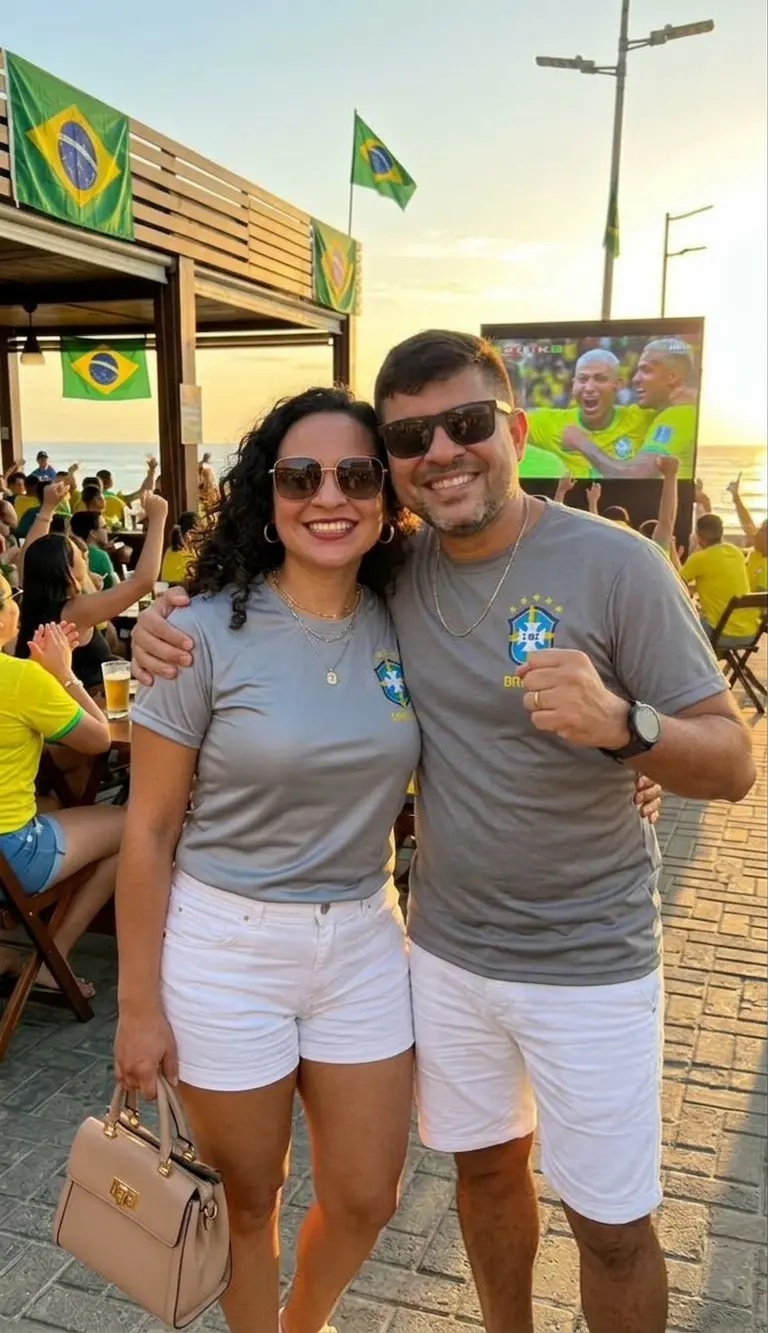 Casal sorrindo vestindo uma blusa do Brasil na cor cinza com escudo e detalhes em amarelo, combinada com shorts branco. Produto da loja Leticy Moda Feminina.