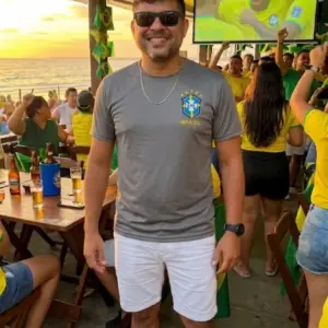 Homem sorrindo vestindo uma blusa do Brasil masculina na cor cinza com escudo e detalhes em amarelo, combinada com shorts branco. Produto da loja Leticy Moda Feminina.